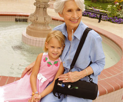 Die Großmutter sitzt mit Ihrer Enkelin am Rand eines Springbrunnens. Die Großmutter hat eine Sauerstoffflasche in einer Tragetasche über der Schulter hängen. - air-be-c Medizintechnik