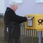 Ein Mann steht an einem Briefkasten und wirft einen Brief ein, er trägt um die rechte Schulter eine abgefüllte Sauerstoffflasche.