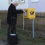 Ein älterer Herr steht an einem Briefkasten und wirft einen Brief ein, der iGo steht neben ihm.