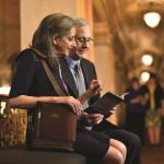 Ein älteres Ehepaar sitzt im Theater Foyer und schaut sich das Programmheft an. Die Frau trägt den SimplyGo mini in der Tragetasche über die Schulter gehängt. - SimplyGo mini