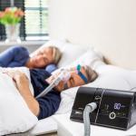 Ein älteres Ehepaar ruht sich aus. Der Mann trägt eine Pillow Maske. Auf dem Nachttisch steht der prisma20C